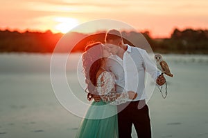 Bride and groom with small owl in hands are standing, huging and kissing on background of sunset.