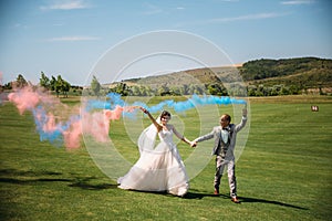 The bride and groom with smoke bombs on a field with green grass. Newlyweds walking outdoors at wedding day. Girl in