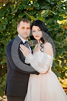 bride and groom stand by a bush with green foliage in the park.
