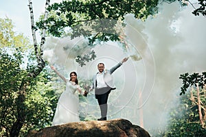 The bride and groom stand on a cliff and hold smoke bombs in their hands in white