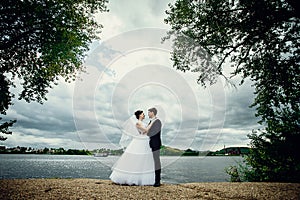 The bride and groom stand on the shore of the pond and look at each other. Beautiful full-length portrait