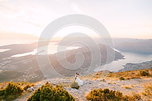The bride and groom stand on the top of Mount Lovcen overlooking the Bay of Kotor near wooden bench, kiss and hug