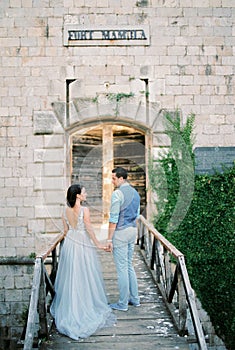 Bride and groom stand on the wooden bridge of Mamula Fort, holding hands. Back view