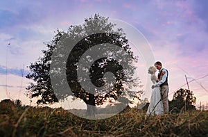 The bride and groom are standing next to a large tree, after the wedding ceremony. Silhouette photo in warm colors.