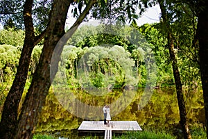 The bride and groom are standing on a wooden pier near the pond