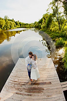 The bride and groom are standing on a wooden pier near the pond
