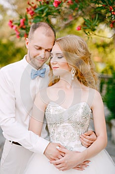 The bride and groom tenderly snuggle against the background of a flowering oleander bush near the ancient church in