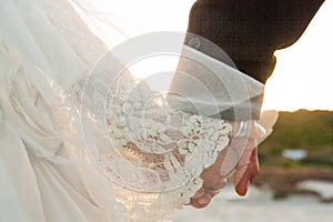 A bride and groom with vintage wedding dress hold hands walking on a beach towards the sunset