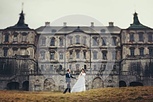 Bride and groom walk along the yellow lawn in the front of an old ruined castle