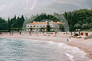 The bride and groom walk holding hands on the pebbled seashore near beautiful villa