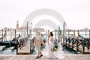 The bride and groom are walking along the gondola pier, holding hand in Venice, near Piazza San Marco, overlooking San