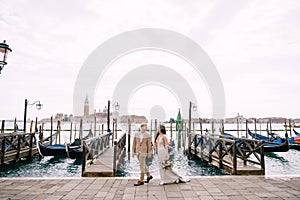The bride and groom are walking along the gondola pier, holding hand in Venice, near Piazza San Marco, overlooking San