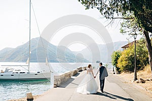 The bride and groom are walking hand in hand on the road by the sea, a white yacht is standing near the shore