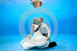 The bride and groom in wedding dresses embrace and kiss underwater at the bottom of a swimming pool