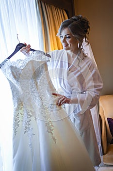 Bride holding wedding dress on a hanger near window and smiling