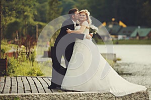 Bride holds groom's hand while he hugs her on the lake's shore