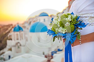Bride holds wedding bouquet in white and green colors and blue decor against the backdrop of the sunset over Santorini, Greece