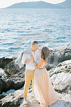 Bride hugs groom by the neck and almost kisses on the rocks by the sea