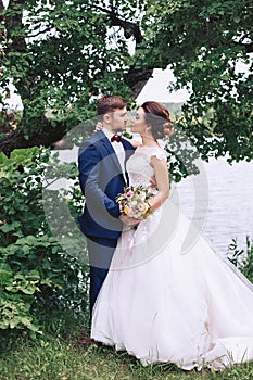 The bride hugs the groom on the river bank under the trees