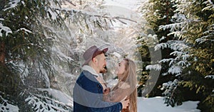 bride laughing hugging kiss groom hat denim jacket under falling snow in forest Christmas tree