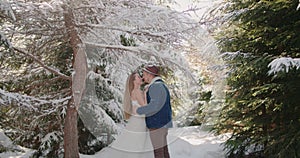 bride laughing hugging kiss groom hat denim jacket under falling snow in forest Christmas tree