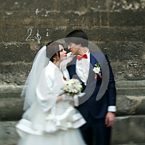 Bride leans on groom`s shoulder standing with him behind a stone