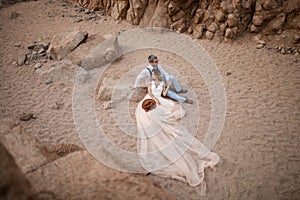 Bride in long dress and groom sit and hold hands in canyon on sand. Top view.