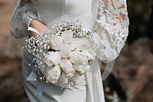 The bride in a long tight white wedding dress holds a beautiful bouquet of white roses in her hands