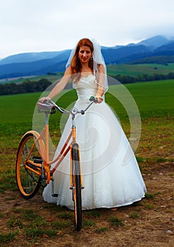 Bride on orange bike in beautiful wedding dress with lace in landscape.