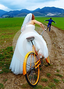 Bride on orange retro bike is chasing after a groom in blue wedding suit with a beer bottle. wedding concept.