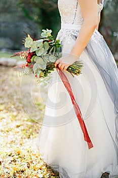 A bride in a pale gray wedding dress holds a bouquet with red peonies, astilba, roses and eringium and red long ribbons