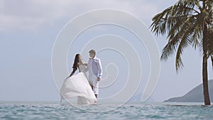 Bride is playing with her long white wedding dress. Young beautiful man and woman hug each other at the beach. Happy