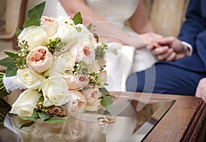 Bride`s bouquet with pink and beige roses,  gold wedding rings on the table, silhouettes of the bride and groom in the distance