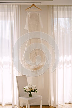 The bride`s dress by the curtains on the window above the chair with a bouquet of flowers.