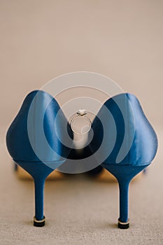 Bride`s engagement ring with a gem between blue high-heeled shoes on the floor on a beige background.