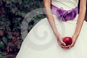 Bride`s hands holding red apple - symbol of love - over white vapory dress and green garden. Vintage style. Close up. Copy-space.