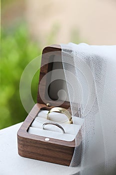 Bride\'s veil drapes over wedding rings in simple wooden box for outside wedding ceremony
