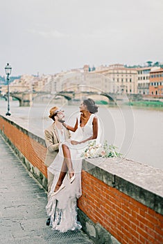 Bride sits on a brick fence next to the groom on the river bank