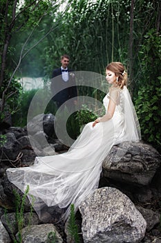 The bride sits on a large stone near the river. A bride stands near the bride