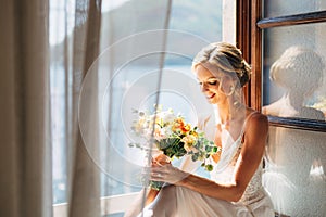 The bride sits at an open window with a sea view and fluttering curtains and holds a wedding bouquet with orange flowers