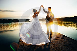 Bride is spinning in a white dress holding hand the groom on the bank of the lake at sunset