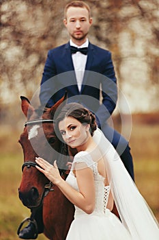 Bride stands behind a horse while groom sits on its back