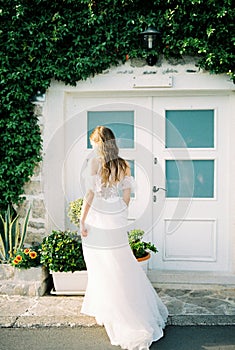 Bride stands in front of the white door of a house overgrown with green ivy. Back view