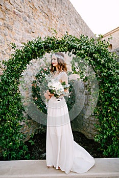 A bride stands at a graceful arch entwined with wild grapes in the old town of Budva and holds a bouquet in her hands