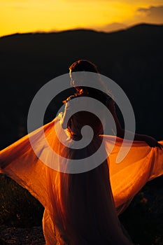 Bride stands holding the hem of the dress on the mountain against the backdrop of the sunset