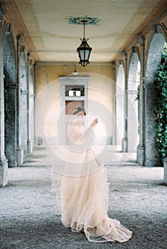 Bride stands on the terrace covering her face with a hem of the dress