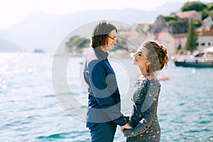 The bride in a stylish grey wedding dress and the groom stand holding hands on the seashore near the old town of Perast