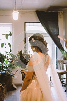 Bride waiting for the big moment. Young bride in beautiful dress holding bouquet of flowers posing near window at home. Wedding