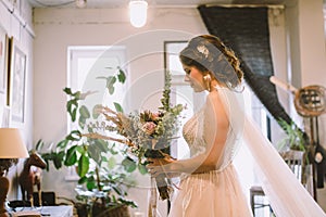 Bride waiting for the big moment. Young bride in beautiful dress holding bouquet of flowers posing near window at home. Wedding