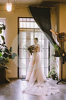 Bride waiting for the big moment. Young bride in beautiful dress holding bouquet of flowers posing near window at home. Wedding